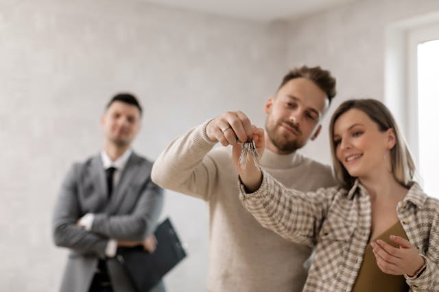 A tenant couple holding the keys to their new home, while a landlord watches in the background, smiling
